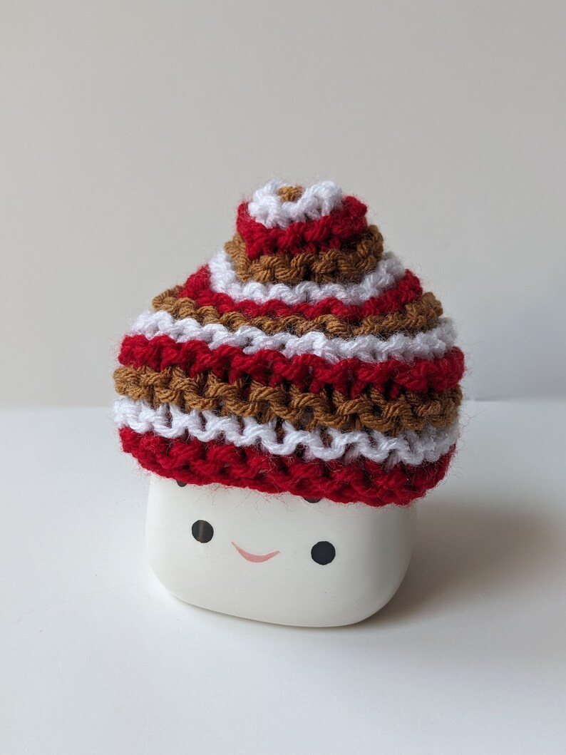 Puede incluir: Una taza de cer&aacute;mica blanca con una cara sonriente, que lleva un gorro de punto con rayas rojas, blancas y marrones.
