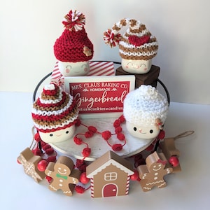 Puede incluir: Una bandeja de tres niveles decorada para Navidad con un tema de pan de jengibre. La bandeja presenta cuatro tazas blancas con gorros tejidos, un letrero que dice "Mrs. Claus Baking Co. Homemade Gingerbread Cakes, Cookies, Candles", y una guirnalda de pompones rojos y hombres de jengibre de madera.