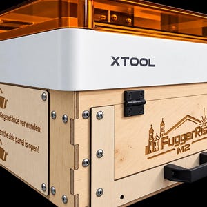 Może przedstawiać: Maszyna laserowa xTool z obudową z jasnego drewna, białą górną częścią i pomarańczową, półprzezroczystą pokrywą. Przedni panel zawiera wygrawerowany napis FuggerRise M2, a boczny panel zawiera teksty i ikony dotyczące bezpieczeństwa użytkowania.