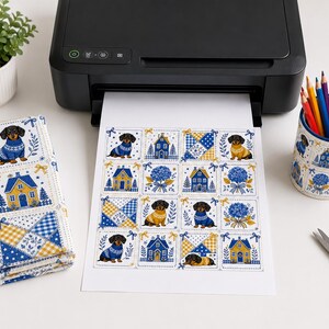 May include: A black printer outputting a sheet of paper with a grid pattern featuring illustrations of dachshunds in sweaters, blue houses, yellow and blue checkered squares, and blue hydrangea bouquets, matching a fabric stack and a pencil holder.
