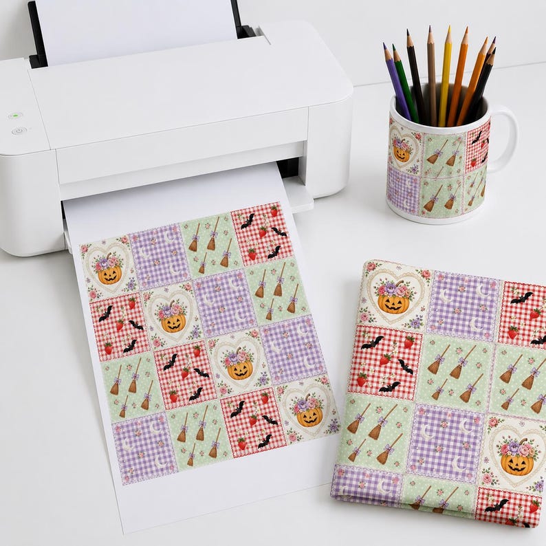 Op de afbeelding: Een witte printer voert een vel papier uit met een herhalend rasterpatroon van pompoenen, heksenbezems, vleermuizen en aardbeien. Een bijpassende keramische mok gevuld met kleurpotloden staat ernaast, samen met een gevouwen vierkant van stof.