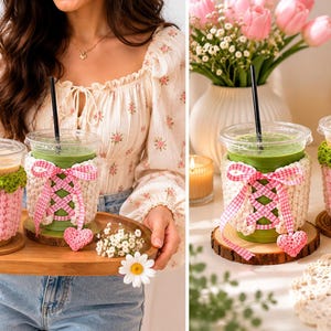 May include: Two clear plastic iced coffee cups featuring handmade crochet sleeves. One sleeve is pink with a white flower, and the other is cream-colored with a pink gingham ribbon lace-up detail and a small dangling pink heart charm.