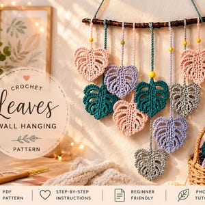 Puede incluir: Un colgante de pared de ganchillo con adornos en forma de hojas en varios colores, incluyendo rosa, verde azulado y lavanda, suspendidos de una rama de madera. La imagen también incluye un ovillo de lana, un ganchillo y una cesta.