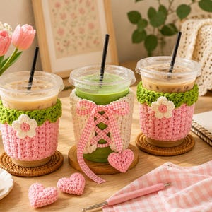 May include: Three clear plastic iced beverage cups with black straws, each featuring a handmade crochet cozy. Two cozies are pink with green scalloped tops and white flower appliqu&eacute;s. The center cozy is cream-colored with a pink gingham ribbon lace-up.