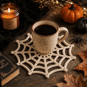 May include: A beige crocheted spider web coaster sits under a speckled ceramic mug filled with black coffee. The coaster features a radial web pattern with interconnected yarn loops, providing a textured base for the drinkware in this setting.