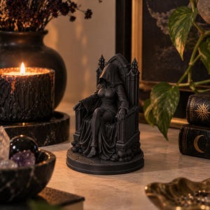 May include: A dark grey figurine of a hooded figure sitting on a high-backed gothic throne. The figure wears a long, flowing robe with draped fabric and is positioned on a circular base with a stone-like texture and visible architectural details.