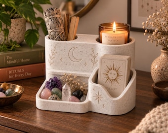 May include: A speckled white multi-compartment organiser featuring engraved moon, star, and leaf motifs. It holds a bundle of sage, wooden sticks, a lit Sandalwood Vanilla candle, a deck of tarot cards with a sun illustration, and various crystals.