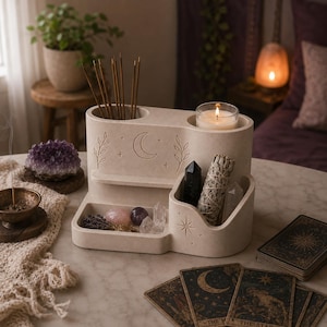 May include: A beige stone-like organizer featuring a crescent moon, star, and leaf engravings. It includes two cylindrical top slots for incense and a candle, a small shelf, and a deep front compartment holding crystals and a sage bundle.