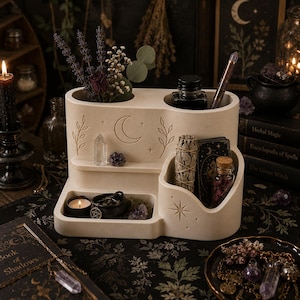 May include: A beige multi-tiered desk organizer featuring engraved celestial motifs including a crescent moon, stars, and leafy branches. The organizer has multiple compartments holding dried flowers, a glass ink bottle, a pen, a sage bundle, and a candle.