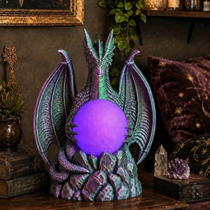 May include: A dragon figurine with iridescent green and purple scales perched on a rocky base. The dragon has large, textured wings and holds a glowing, spherical purple orb between its front claws. The sculpture features intricate detailing.