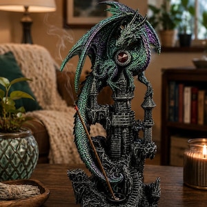 May include: A green and purple dragon figurine perched atop a dark grey stone castle tower. The dragon features a prominent red circular gem on its chest. A thin incense stick is inserted into a small holder at the base, emitting a trail of smoke.