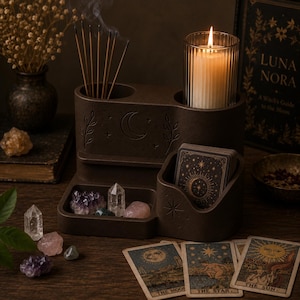 May include: A dark brown multi-functional organizer featuring a crescent moon and leaf engraving. It includes a cylindrical holder for incense sticks, a ribbed glass candle holder with a lit candle, and a compartment for a deck of tarot cards.