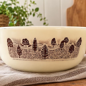 May include: A cream-colored glass mixing bowl with two side handles, featuring a dark brown printed graphic of a forest landscape with various trees and rolling hills encircling the exterior of the round, wide-rimmed kitchen vessel.