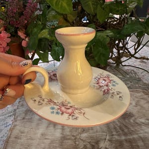 Peut inclure: Un bougeoir en céramique de couleur crème avec une anse, orné d'un motif floral délicat dans des tons rose et bleu. L'objet présente une finition émaillée craquelée et un fin liseré rose autour de l'ouverture supérieure et du bord de la base.
