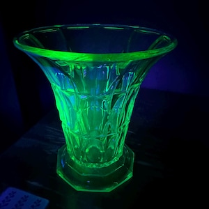 Puede incluir: Un jarrón de vidrio verde neón vibrante y brillante que presenta un patrón geométrico facetado en todo su cuerpo. El jarrón tiene un borde ancho y acampanado y una base hexagonal resistente, iluminado bajo luz ultravioleta intensa.