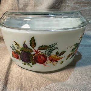 Peut inclure: Une cocotte en verre opalin blanc avec un couvercle en verre transparent. L'extérieur présente une illustration botanique colorée de divers légumes, dont une aubergine violette, une betterave rouge, une carotte et des herbes vertes.