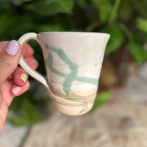 Peut inclure: Une tasse en céramique avec une partie supérieure émaillée blanche ornée d'un motif de lignes abstraites vert clair. La partie inférieure de la tasse n'est pas émaillée, révélant une texture d'argile beige naturelle avec des stries horizontales.