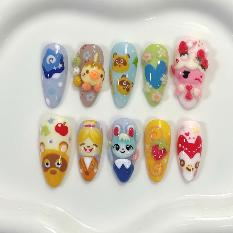May include: A set of ten almond-shaped acrylic nails, each uniquely decorated with colorful, cartoon-style designs. The designs include characters, patterns, and 3D elements in various colors, including blue, yellow, pink, and green.