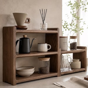 May include: A two-tiered wooden shelf unit made of dark walnut wood, featuring rounded edges. The shelves hold a ceramic coffee dripper, a black gooseneck kettle, a white mug, two stacked bowls, and a glass jar filled with coffee beans.