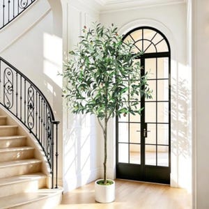 May include: A tall artificial olive tree with slender green leaves and small dark olives, featuring a single thin trunk planted in a simple white cylindrical pot, positioned upright on a light-colored floor in a bright indoor space.