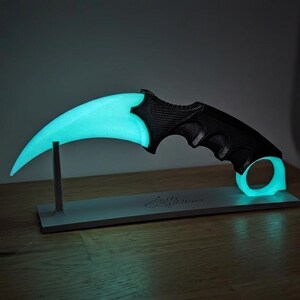 Pode incluir: Uma faca karambit preta e verde-azulado com uma lâmina curva e um anel para o dedo. A lâmina e o anel brilham com uma luz verde-azulado. A pega é preta com uma pega texturizada. A faca é exibida em um suporte cinza.