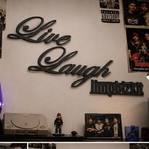 Puede incluir: Un letrero negro montado en la pared con las palabras Live Laugh y el nombre de la banda Limp Bizkit en fuente cursiva y de bloque. El letrero se muestra sobre una configuración de dormitorio con un televisor, una lámpara de lava y pósteres.