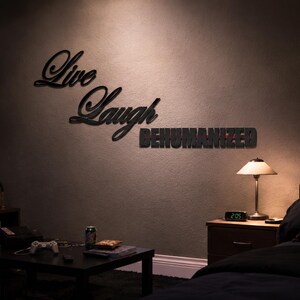 Puede incluir: Un letrero de pared negro en letra cursiva que dice Live Laugh y un letrero en letras de bloque que dice DEHUMANIZED. El texto se muestra en una pared oscura en una habitación que contiene un televisor, una mesa de centro, una cama y una lámpara.