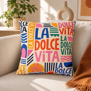 May include: A square throw pillow with a cream-colored background featuring a repeating graphic pattern of the text La Dolce Vita in various bold colors including blue, orange, pink, and green, interspersed with abstract shapes and wavy lines.