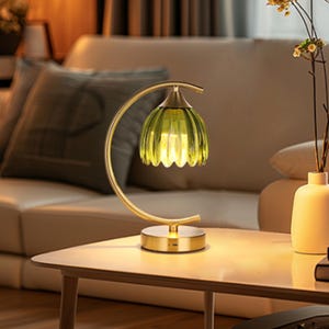 May include: A decorative table lamp with a gold-colored curved frame and base. The lamp features a green, ribbed glass shade. The lamp is on a light-colored wooden table in a living room setting.