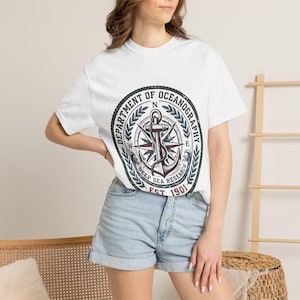 以下が含まれることがあります： 胸元に円形のグラフィックがプリントされた白い半袖Tシャツ。デザインにはコンパスローズ、錨、月桂樹の葉が含まれており、「DEPARTMENT OF OCEANOGRAPHY DEEP SEA RESEARCH EST 1901」という文字が暗い色で描かれています。