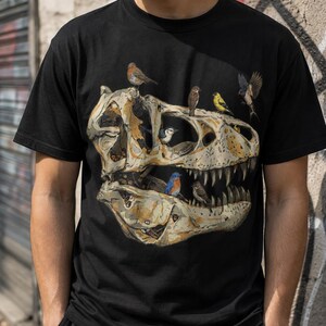 May include: A black short-sleeved t-shirt featuring a large graphic print of a beige dinosaur skull. The skull is decorated with several small, colourful birds perched on and inside the bone structure, including a blue bird and a yellow bird.