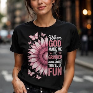 May include: Black t-shirt featuring a pink sunflower design with butterflies and the text "When God Made Me He Grinned and Said This'll Be Fun". The shirt is worn by a person in black jeans.