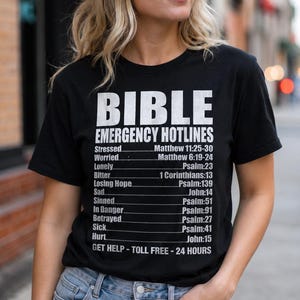 May include: Black t-shirt with white text. The shirt reads "BIBLE EMERGENCY HOTLINES" with a list of emotional states and corresponding Bible verses. The wearer is also wearing blue jeans.