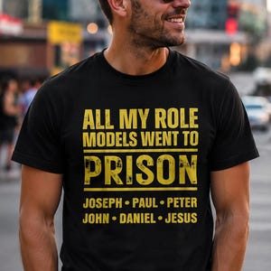 May include: Black t-shirt with the text "ALL MY ROLE MODELS WENT TO PRISON" in yellow. Below the text are the names Joseph, Paul, Peter, John, Daniel, and Jesus. The shirt is worn by a person wearing sunglasses.