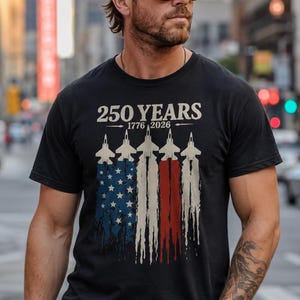 Könnte beinhalten: Schwarzes T-Shirt mit patriotischem Design. Das Shirt zeigt den Text "250 YEARS 1776-2026" über einer Grafik von Jets, die die amerikanische Flagge bilden. Die Jets hinterlassen Spuren in Rot, Weiß und Blau.