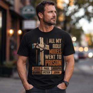 May include: A black short-sleeved t-shirt featuring a graphic of a wooden cross with a crown of thorns. The text reads: ALL MY ROLE MODELS WENT TO PRISON. Below, a grid lists the names: JESUS, PAUL, JOSEPH, DANIEL, PETER, and JOHN.