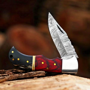 May include: A folding pocket knife featuring a Damascus steel blade with a wavy pattern. The handle is constructed from a combination of black horn and red wood, accented with small brass pins and a central brass spacer ring near the bolster.