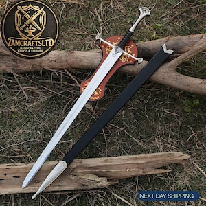 May include: A silver-toned metal sword with a black wrapped handle, a crossguard, and a pointed blade featuring engraved text. The sword rests on a brown wooden plaque with gold floral patterns, accompanied by a black scabbard with silver metal accents.