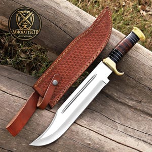 May include: A fixed-blade knife with a polished steel blade, a brass guard, and a handle featuring alternating dark wood and black rings. The knife includes a textured brown leather sheath with a snap closure and a circular logo reading ZAMCRAFTSLTD.