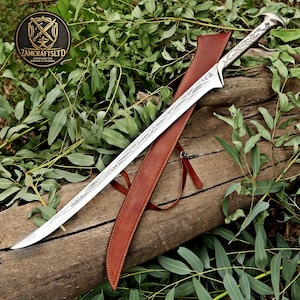 May include: A silver-coloured metal sword with an elongated, curved blade featuring intricate etched vine-like patterns. The hilt has a matching decorative design and a pommel. A brown leather scabbard with stitched edges and leather straps lies beneath.