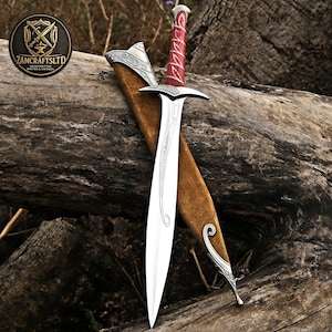 May include: A silver-toned metal sword with a pointed blade featuring etched scrollwork, a crossguard, and a red handle wrapped in silver wire. The sword is accompanied by a brown suede-like scabbard with silver metal fittings at the top and tip.