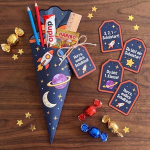 May include: A dark blue cone-shaped gift bag decorated with space-themed illustrations including a rocket, planet, and moon. It contains a wooden ruler, a Duplo chocolate bar, a Haribo candy pack, and two pencils. Four matching gift tags are included.