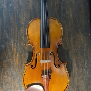 May include: A detailed, antique-style violin with a warm, golden-brown finish. The instrument features a dark fingerboard, strings, and a reddish-brown chin rest. The body has decorative edging and f-holes, showcasing the craftsmanship of a stringed instrument.