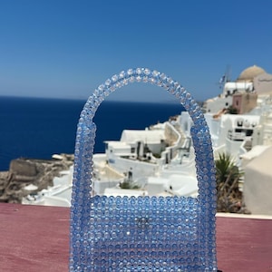 Può includere: Una piccola borsa realizzata interamente con perline traslucide azzurre e trasparenti. La borsa presenta un corpo rettangolare con un manico arrotondato, il tutto creato con un motivo a griglia ripetuto di perline sferiche.
