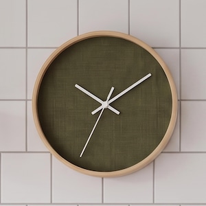 May include: A round wall clock featuring a light-coloured wooden frame and a dark olive green fabric face with a textured weave. The clock has three thin white hands positioned at the centre, with no numbers or markings on the clock face.