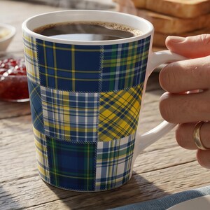 May include: A ceramic mug featuring a patchwork design of various tartan patterns in shades of navy blue, bright yellow, forest green, and white. The mug has a white interior and a white handle, and it is filled with a dark brown liquid beverage.