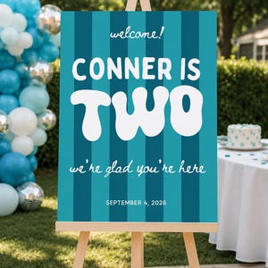 Puede incluir: Un letrero de bienvenida rectangular sobre un caballete de madera con rayas verticales en azul cerceta y azul oscuro. El texto dice: welcome! CONNER IS TWO we're glad you're here SEPTEMBER 4, 2026, con letras blancas en fuentes cursivas y negritas.