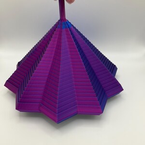 Könnte beinhalten: Ein geometrisches, mehrschichtiges Objekt in Lila- und Blautönen wird von einer Hand gehalten. Das Objekt hat eine sternartige Form mit einem Griff. Der Farbverlauf geht von tiefem Lila zu helleren Tönen mit blauen Akzenten über.