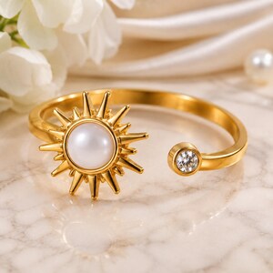 May include: A gold-toned open ring featuring a central sunburst motif with a smooth white pearl-like cabochon at its core. The opposite end of the band is finished with a single small, clear, round-cut gemstone set in a gold-toned bezel.