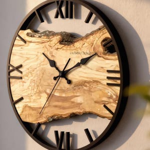 Könnte beinhalten: Eine runde Wanduhr mit schwarzem Rahmen und römischen Ziffern. Das Zifferblatt besteht aus hellem Holz mit einem einzigartigen Maserungsmuster. Schwarze Zeiger zeigen die Uhrzeit an. Die Uhr ist ein dekoratives Wohnaccessoire.